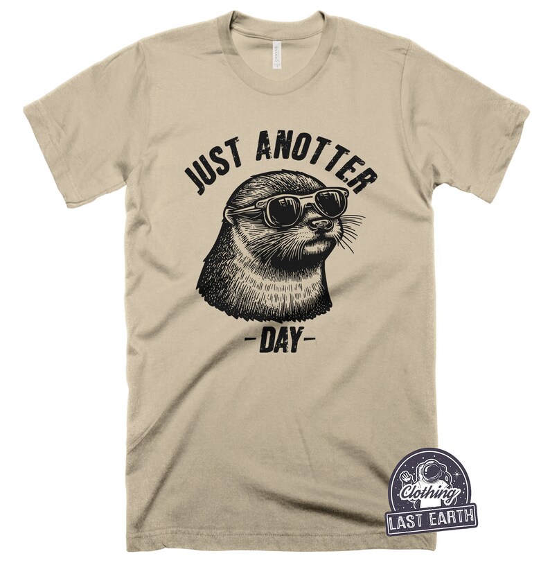 May include: A tan t-shirt with a black graphic of an otter wearing sunglasses and the text "Just Anotter Day".