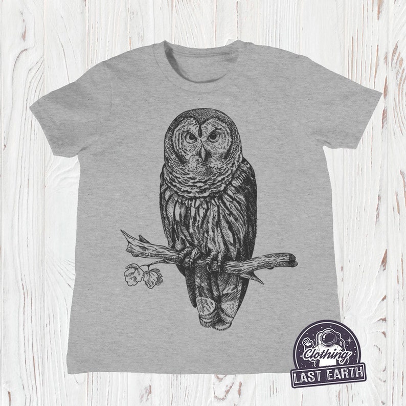 May include: A gray t-shirt with a black and white illustration of an owl perched on a branch. The owl has large, round eyes and detailed feathers. The t-shirt has the text "Clothing Last Earth" in a circle with a spaceman graphic.