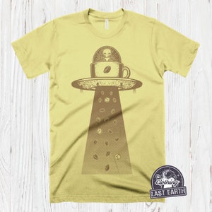 May include: A yellow t-shirt with a graphic of a UFO shaped like a coffee cup with an alien inside. Coffee beans are falling from the UFO. The text "Clothing Last Earth" is on a patch in the bottom right corner.
