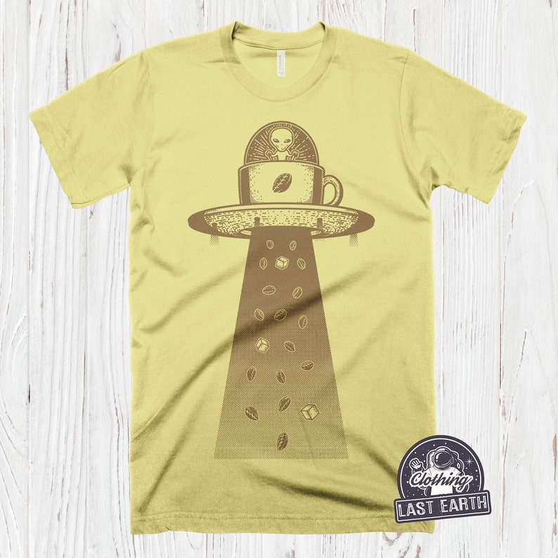 May include: A yellow t-shirt with a graphic of a UFO shaped like a coffee cup with an alien inside. Coffee beans are falling from the UFO. The text "Clothing Last Earth" is on a patch in the bottom right corner.