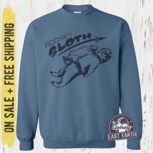 Könnte beinhalten: Ein blaues Sweatshirt mit einem schwarzen Aufdruck eines Faultiers, das einen Raketenrucksack trägt, und dem Text "Rocket Sloth".