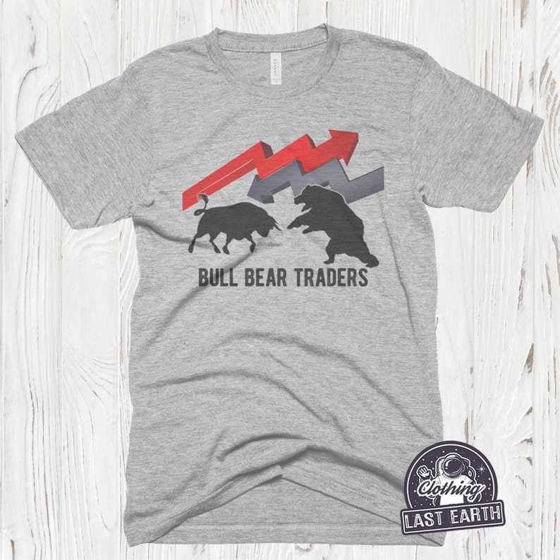 Puede incluir: Una camiseta gris con un gr&aacute;fico de un toro y un oso enfrentados. El toro es rojo y el oso es negro. Por encima de ellos hay dos flechas rojas y grises que apuntan hacia arriba. El texto "BULL BEAR TRADERS" est&aacute; impreso debajo del gr&aacute;fico.