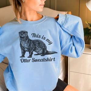 May include: A light blue sweatshirt with a black and white illustration of an otter. The text "This is my Otter Sweatshirt" is printed on the front of the sweatshirt.