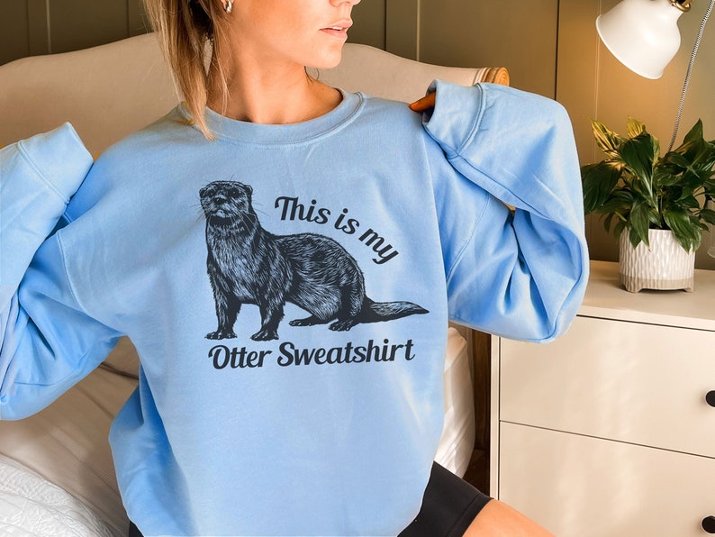 May include: A light blue sweatshirt with a black and white illustration of an otter. The text "This is my Otter Sweatshirt" is printed on the front of the sweatshirt.