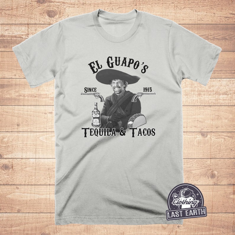 K&ouml;nnte beinhalten: Ein graues T-Shirt mit einem schwarz-wei&szlig;en Grafikdesign. Die Grafik zeigt einen Mann mit einem Sombrero, der eine Flasche und zwei Pistolen h&auml;lt. Der Text auf dem Shirt lautet "El Guapo's Since 1915 Tequila & Tacos".