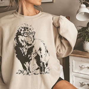 Può includere: Una felpa beige con una stampa grafica in bianco e nero di un leone con gli occhiali. Il leone è in piedi su un pezzo di erba.