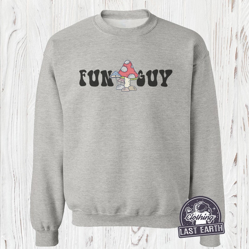 K&ouml;nnte beinhalten: Ein graues Sweatshirt mit dem Text "FUN GUY" in schwarzen Buchstaben. Der Text ist mittig auf dem Sweatshirt platziert und wird von einem Cartoon-Bild von drei Pilzen begleitet, einem roten mit wei&szlig;en Punkten, einem blauen und einem braunen.