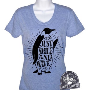Pu&ograve; includere: Una maglietta blu chiaro con un grafico di pinguino nero. Il pinguino tiene una tavola da surf con il testo "Just Smile And Wave" su di essa. La maglietta ha un collo a V e maniche corte.