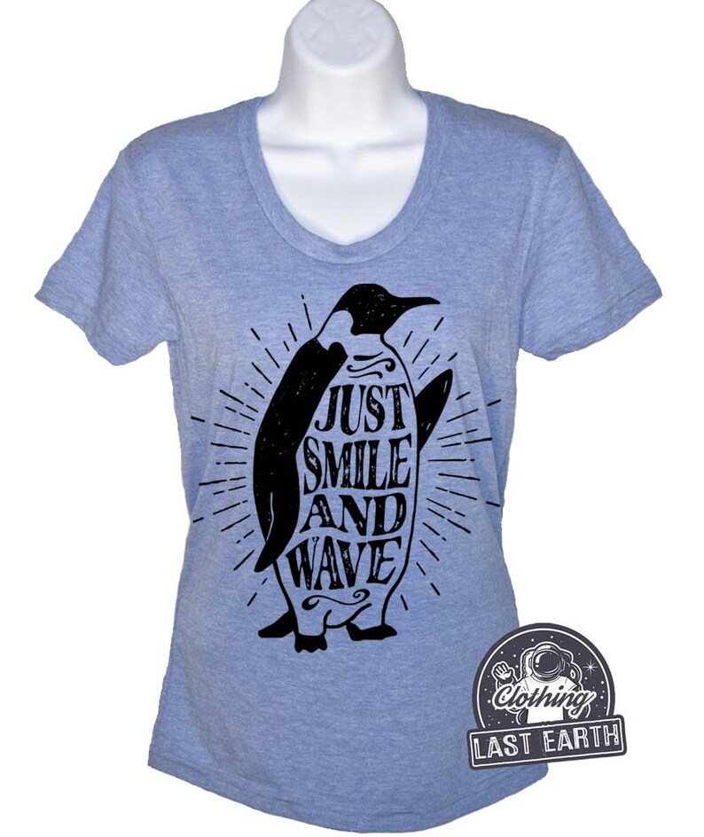 Pu&ograve; includere: Una maglietta blu chiaro con un grafico di pinguino nero. Il pinguino tiene una tavola da surf con il testo "Just Smile And Wave" su di essa. La maglietta ha un collo a V e maniche corte.