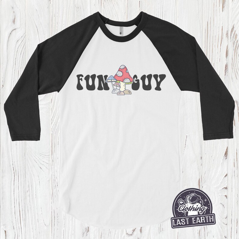 K&ouml;nnte beinhalten: Ein wei&szlig;es und schwarzes Baseball-T-Shirt mit einem bunten Pilz-Motiv und dem Text "FUN GUY" in einer Retro-Schrift.
