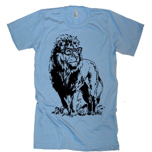 May include: A light blue t-shirt featuring a black and white illustration of a lion wearing glasses. The lion is standing on a grassy surface.
