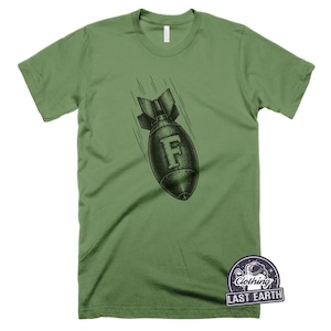 May include: An olive green short-sleeve t-shirt featuring a black graphic of a falling bomb with the letter F centered on its side. The graphic includes motion lines indicating downward movement. A small logo in the corner reads Clothing LAST EARTH.