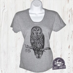 May include: Gray t-shirt with a black and white illustration of an owl perched on a branch. The text "Clothing Last Earth" is printed on a small patch in the lower right corner of the shirt.