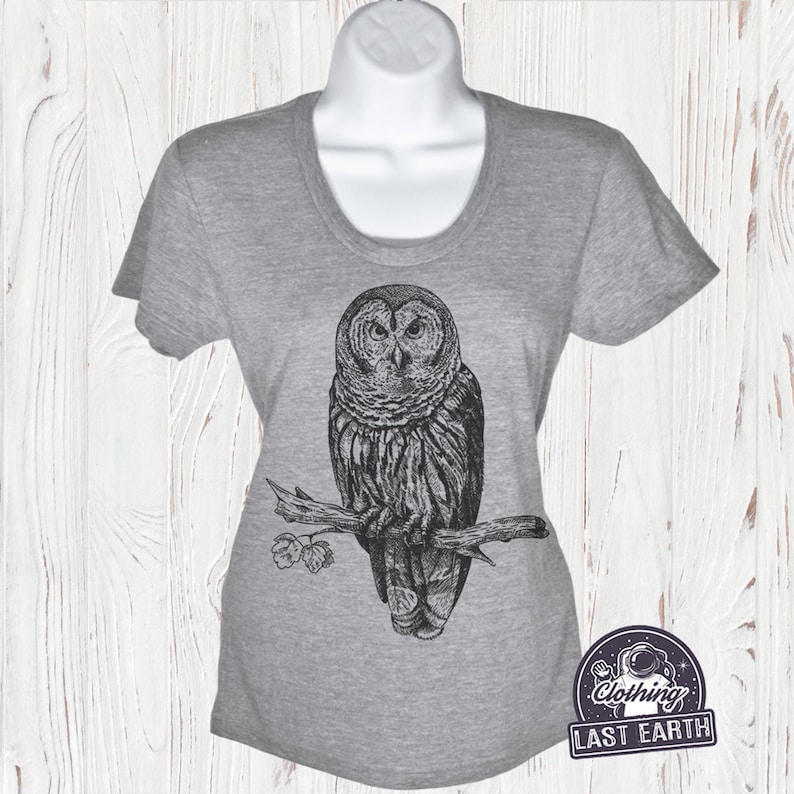 May include: Gray t-shirt with a black and white illustration of an owl perched on a branch. The text "Clothing Last Earth" is printed on a small patch in the lower right corner of the shirt.