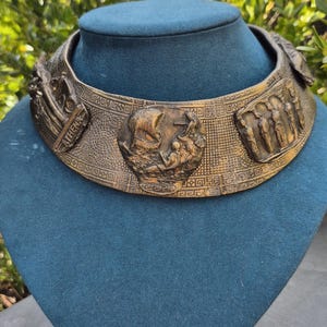 Op de afbeelding: Een brede choker-halsketting van bronskleurig metaal met reliëfscènes en figuren. Het oppervlak toont ingewikkelde geometrische randpatronen, een centraal rond medaillon met een schip en een rechthoekige plaquette met het opschrift ATHENA.