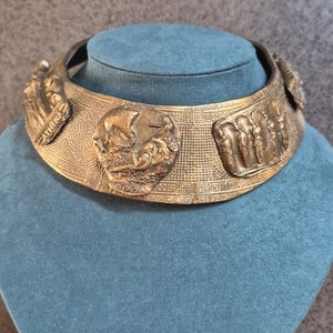 Könnte beinhalten: Ein breites, starres Halsband in Bronzefarbe mit geprägten Reliefplatten. Das zentrale Design zeigt ein Schiff und Figuren, wobei das Wort ATHENA auf einer Seite eingraviert ist. Die Oberfläche ist mit einem geometrischen Muster texturiert.