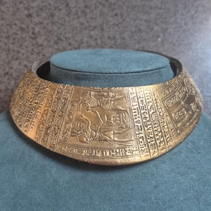 Könnte beinhalten: Ein breites, starres goldfarbenes Halsband mit geprägten Hieroglyphen im ägyptischen Stil und einem zentralen Relief, das Figuren im Profil zeigt. Die metallische Oberfläche hat ein strukturiertes, gealtertes Finish mit detaillierten Gravuren.