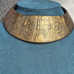 May include: A wide, bronze-colored collar necklace with intricate embossed designs. The necklace features detailed patterns and symbols, possibly of ancient origin, and is displayed on a blue surface.