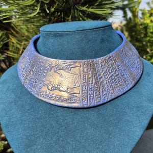 Könnte beinhalten: Ein breites, starres Halsband mit einer blauen Basis und goldfarbenen Prägedetails. Die Oberfläche ist mit komplizierten Mustern verziert, die an altägyptische Hieroglyphen und Figuren erinnern und ein strukturiertes, metallisches Aussehen erzeugen.