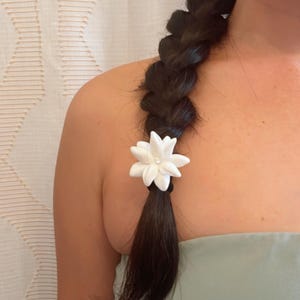 Op de afbeelding: Een donkerbruin gevlochten kapsel met een wit bloemvormig haaraccessoire. De bloem heeft de vorm van een ster met een kleine parel in het midden. Het accessoire is bevestigd aan een zwarte elastiek.