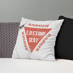 May include: White throw pillow with a red triangle and the words "DANGER EJECTION SEAT" in red and white. The pillow has a riveted metal design, and the word "DANGER" is repeated around the triangle.
