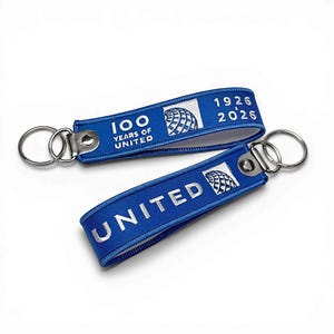 Puede incluir: Dos llaveros azules con anillos de metal plateado. Los llaveros tienen texto bordado blanco y un logotipo. Un llavero dice "100 YEARS OF UNITED 1926 2026". El otro llavero dice "UNITED".