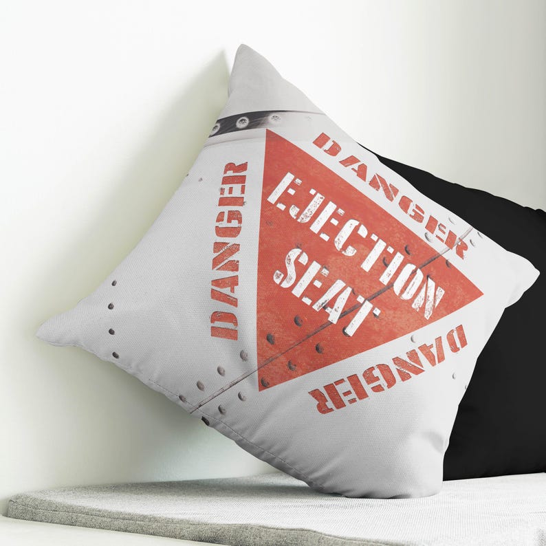 May include: A square decorative pillow with a white background and a red triangle in the center. The triangle has the words "EJECTION SEAT" in white, with "DANGER" in red around the edges. The pillow has a riveted metal design.