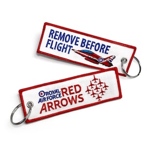 May include: Two rectangular embroidered keyrings with red borders and silver rings. One reads "REMOVE BEFORE FLIGHT" with a red airplane. The other says "ROYAL AIR FORCE RED ARROWS" with a formation of red aircraft.