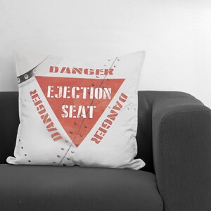 May include: White square pillow with a red triangle design that reads "DANGER EJECTION SEAT" in bold, white letters. The pillow has a vintage, distressed look with rivets and a metal plate design.