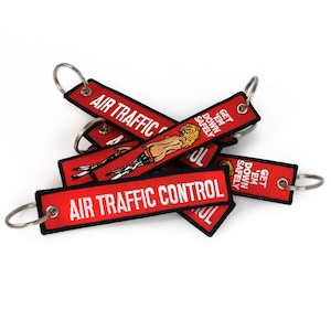 May include: A pile of red keychains with the text "AIR TRAFFIC CONTROL" in white. Some keychains have an illustration of a woman and the text "GET 'EM DOWN SAFELY". Each keychain has a silver metal ring.