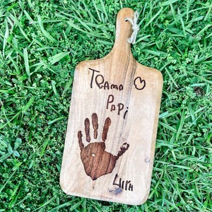Peut inclure: Planche à découper en bois avec une poignée et un motif d'empreinte de main. La planche porte les mots "Te amo Papi" et le nom "Lira" gravés dans le bois. Une boucle en ficelle naturelle est attachée à la poignée.
