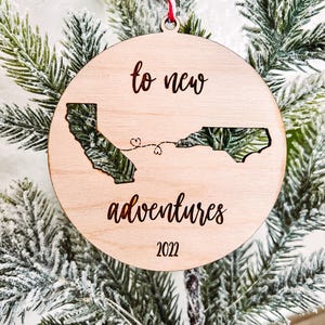 May include: A round wooden Christmas ornament with the text "to new adventures 2022". The ornament features cut-out shapes of two states connected by a dotted line, hanging on a Christmas tree.