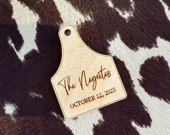 May include: A light brown wooden tag engraved with the words "The Negretes" and "OCTOBER 12, 2025". The tag has a hole at the top. The background features a brown and white cowhide pattern.