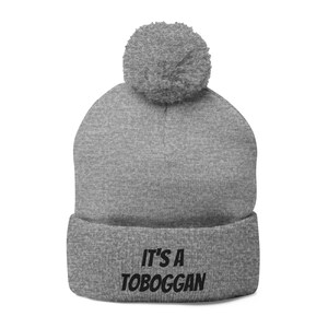 May include: A gray beanie with a matching pom-pom on top. The folded brim of the hat displays the text "IT'S A TOBOGGAN" in black, a playful statement. This winter hat is designed for warmth and style.