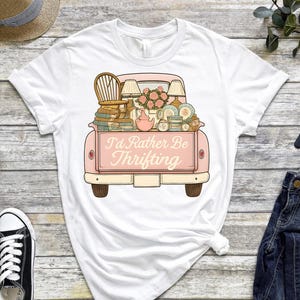 Op de afbeelding: Wit T-shirt met een roze vintage truck afbeelding, vol met tweedehands vondsten. De truck heeft de tekst "I'd Rather Be Thrifting". Het shirt is gestyled met een jeans en sneakers.