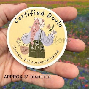 K&ouml;nnte beinhalten: Ein runder Aufkleber mit hellgelbem Hintergrund, der eine Cartoon-Illustration einer Person mit Blumen zeigt. Der Aufkleber tr&auml;gt die Aufschrift "Certified Doula" und "Crunchy but evidence-based". Der Aufkleber hat einen Durchmesser von ca. 7,6 cm.