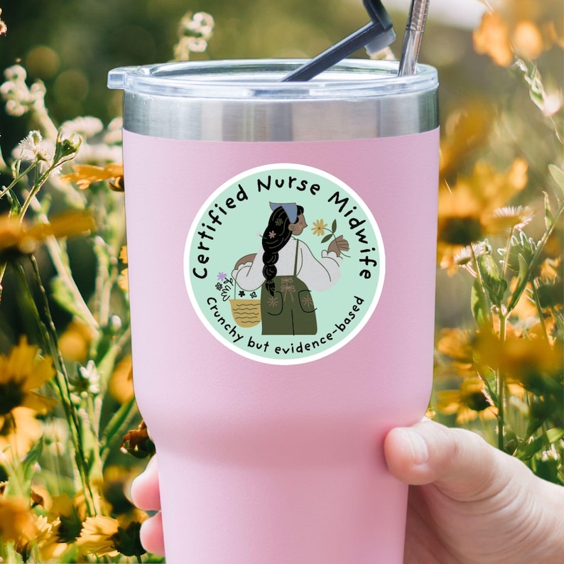 K&ouml;nnte beinhalten: Ein rosa Becher mit einem runden Aufkleber mit einer Cartoon-Illustration einer Hebamme. Der Aufkleber lautet "Certified Nurse Midwife" und "Crunchy but evidence-based". Die Illustration zeigt eine Frau, die Blumen und einen Korb h&auml;lt.