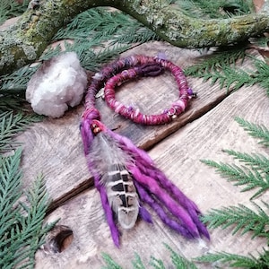 Peut inclure: Extension de dreadlocks en laine violette et rose avec une plume brun et blanc attachée. L'extension est enroulée autour d'une surface en bois.