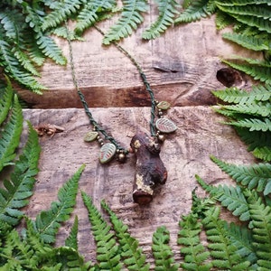 May include: A green and brown necklace with a small wooden branch pendant and green leaf charms. The necklace is displayed on a wooden surface with green ferns.