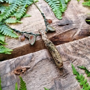 Könnte beinhalten: Eine grüne und braune Halskette mit einem Zweig-Anhänger und Blatt-Anhängern. Die Halskette ist auf einer Holzoberfläche mit grünen Farnen fotografiert.