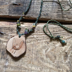 May include: A green cord necklace with a wooden pendant featuring a burnt image of a maple seed. The necklace is adorned with various colored gemstones.