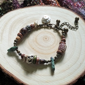 May include: A brown, white, and green beaded bracelet with a white ceramic charm and a metal chain. The bracelet is on a wooden surface.