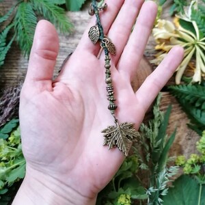 Peut inclure: Un collier pendentif feuille de bronze avec un cordon vert, de petites cloches et des accents de feuilles. Le collier est tenu dans une main, avec un fond en bois et de la verdure.