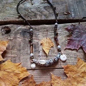 May include: A brown and white beaded necklace with seashells and a copper accent. The necklace is lying on a wooden surface with autumn leaves.