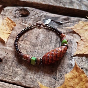 May include: A brown wooden bracelet with green and brown beads, including a large brown pine cone bead. The bracelet is on a wooden surface with fall leaves.