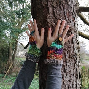 Può includere: Un paio di guanti senza dita colorati con un bordo peloso e un bottone di legno su ogni guanto. I guanti sono realizzati con una varietà di filati in tonalità di verde, blu, viola, arancione e marrone.