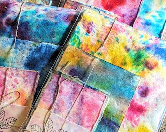 Puede incluir: Una pila de hojas de papel de colores, teñidas a mano, atadas con un cordel. El papel tiene un efecto acuarela con tonos de azul, verde, amarillo, rosa y morado. El papel está decorado con un simple dibujo de hojas.