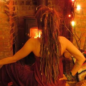 May include: A person with long, colorful dreadlocks is sitting in front of a fireplace. They are wearing a burgundy dress and have a bracelet on their wrist.