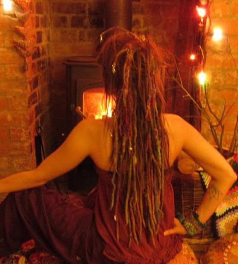 May include: A person with long, colorful dreadlocks is sitting in front of a fireplace. They are wearing a burgundy dress and have a bracelet on their wrist.