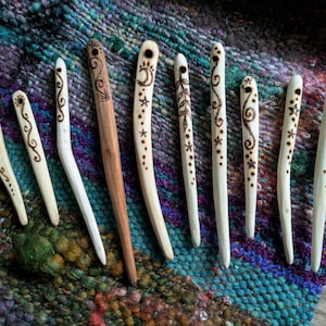 May include: A selection of handcrafted wooden hair sticks, featuring individual burned designs. The sticks range in colour from pale beige to brown, with patterns such as swirls, stars, and botanical motifs. They are displayed on a colourful woven textile.